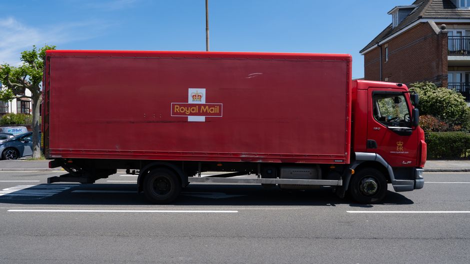 A red Royal Mail delivery truck parked on the side of a suburban street with residential buildings and trees in the background. The truck features a large, enclosed cargo area with the Royal Mail logo and crown symbol on its side. The vehicle is positioned parallel to the sidewalk, with the driver’s cab at the front, and appears to be stationary during the day with clear, bright weather. The street has marked parking bays and a few other vehicles are visible in the background, including a blue car. The scene is well-lit with natural sunlight, emphasizing the clean and orderly environment typical of urban and suburban areas, supporting the outdoor logistics operations associated with home relocation and furniture transportation services, as offered by Removal Companies Enfield.