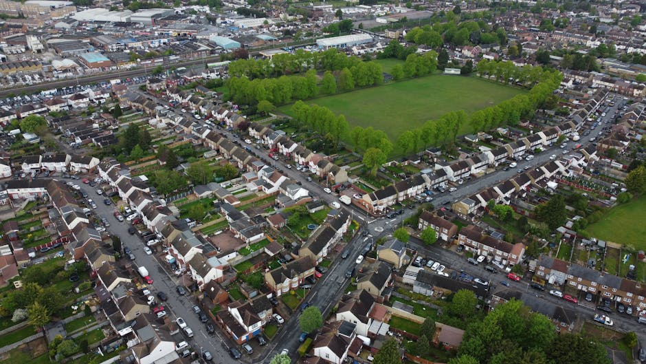 An aerial view of a residential neighbourhood in Enfield showing a dense arrangement of terraced and semi-detached houses with small front gardens, driveways, and parked cars along the streets. In the background, there is a large, rectangular green park surrounded by a line of evenly spaced trees, providing open space within the urban area. The scene captures the landscape during daytime under natural lighting, illustrating the typical environment where home relocation and furniture transport services by Removal Companies Enfield may be required, including the process of packing, loading, and moving household items through streets and onto nearby vehicles or vans parked along the roads.