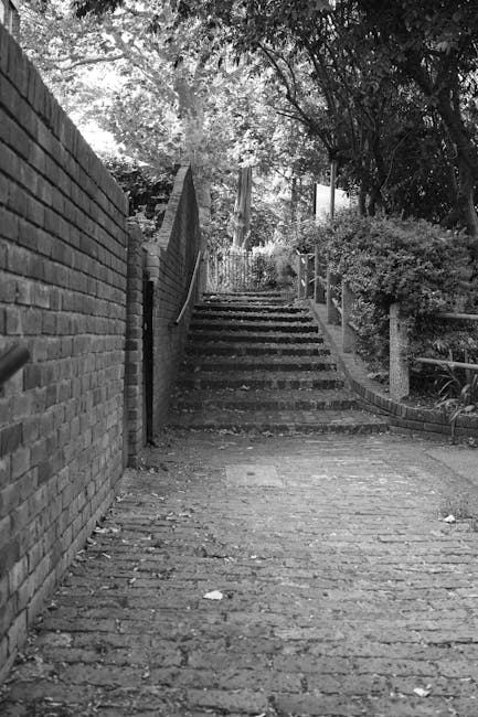 A grayscale image depicting a narrow outdoor street with brick paving. On the left side, a tall brick wall runs alongside the street, while on the right, a low metal railing separates the pathway from