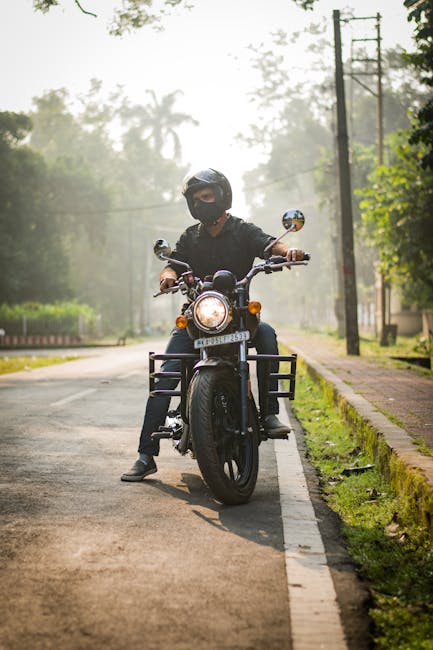A person wearing a black helmet and protective riding gear is seated on a black motorcycle on a suburban street. The motorcycle is positioned in the centre of the lane, with the rider facing forward and the headlight illuminated. The street is lined with grass verges and a paved sidewalk on the right, with a row of utility poles and trees in the background, creating a slightly misty or hazy atmosphere. This scene appears to be outdoors during daytime with soft, natural lighting. The image reflects an element of transport, which could relate to home relocation or moving logistics, consistent with house removals and furniture transport themes supported by Removal Companies Enfield.