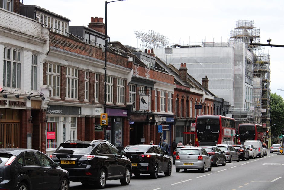 A street scene showing a row of traditional brick terraced houses with large windows, some of which have scaffolding and construction netting, indicating building work or renovations ongoing. Parked along the curb are various cars, including a black Mercedes SUV, a silver hatchback, and other vehicles. There are two red double-decker buses, numbered 343 and 42, positioned on the road, with passengers boarding or alighting. The pavement in front of the houses features pedestrians, some of whom are walking near the entrance doors. In front of the shops, there are shopfronts with large display windows and signs for local businesses. The scene is lit by natural daylight, with an overcast sky, suggesting a typical day involving home relocation activities, such as furniture transport or packing, in the Enfield area. The image is associated with house removals and moving services provided by Removal Companies Enfield, emphasizing the urban environment and city logistics.