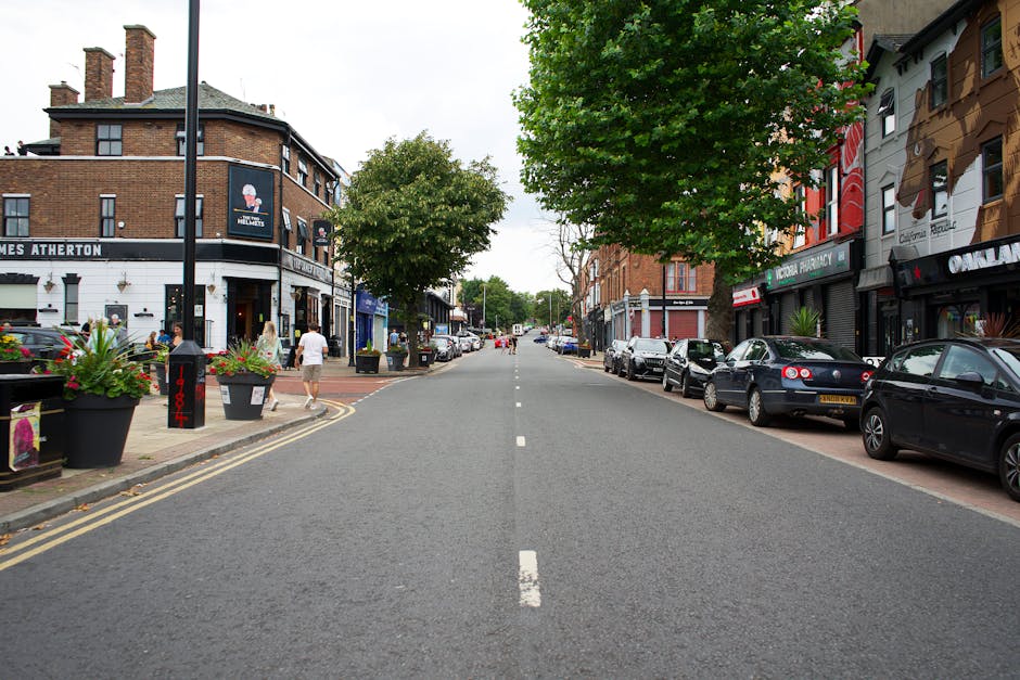 A person wearing a black helmet and protective riding gear is seated on a black motorcycle on a suburban street. The motorcycle is positioned in the centre of the lane, with the rider facing forward and the headlight illuminated. The street is lined with grass verges and a paved sidewalk on the right, with a row of utility poles and trees in the background, creating a slightly misty or hazy atmosphere. This scene appears to be outdoors during daytime with soft, natural lighting. The image reflects an element of transport, which could relate to home relocation or moving logistics, consistent with house removals and furniture transport themes supported by Removal Companies Enfield.
