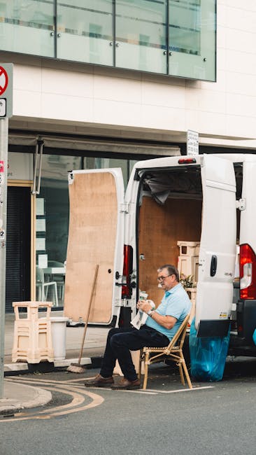 A man is seated on a wooden chair beside a white van parked on a city street, taking a break during a home relocation process. The van’s rear doors are open, revealing a spacious interior filled with various cardboard boxes, some wrapped in plastic and others padded with fabric or packing material. Nearby, a large wooden panel is propped upright against the van, supported by a smaller piece of furniture. The scene shows the man wearing glasses and casual clothing, holding a paper or tablet, with the urban setting of modern office and residential buildings in the background, including a large glass facade and signage indicating a no-parking zone. This snapshot captures the loading process of furniture transport and packing materials involved in professional removals, reflecting a typical move in a densely built area, as undertaken by companies like Removal Companies Enfield, which specialise in house removals and logistical support for tight driveways.