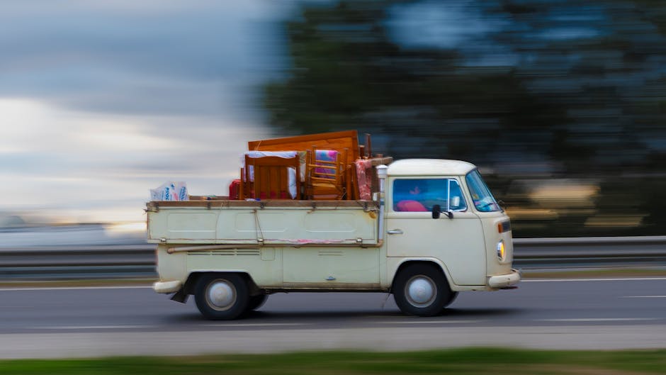 A small vintage white van with a boxy shape and rounded edges is shown moving along a multi-lane road during daytime, with motion blur in the background indicating speed. The van's cargo area is filled with various household furniture and boxes, including a wooden dining chair, a wooden cabinet, cardboard boxes, and plastic-wrapped items, all stacked and secured on the van's flatbed. The scene suggests a house removal or furniture transport scenario, associated with relocation services such as those offered by Removal Companies Enfield, particularly when navigating tight drives or urban access challenges. The lighting is natural, and the environment appears to be suburban with a blurred background of trees and sky, emphasizing the vehicle's motion and the logistical aspects of home relocation involving packing and loading processes.