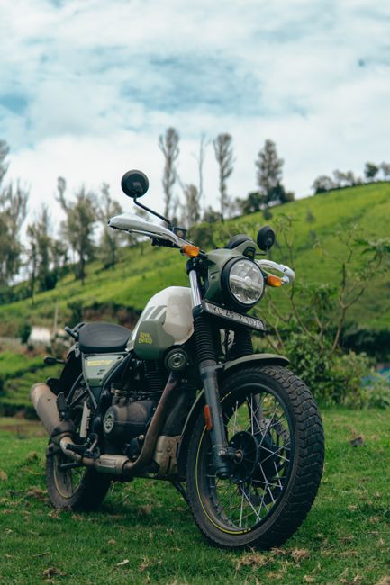A black and white motorcycle with a modern design is parked on a grassy area outdoors, positioned facing slightly to the left. It features a round headlamp, black handlebars with rearview mirrors, and a black seat with a small storage compartment. The motorcycle has visible front and rear wheels with alloy rims, and a black exhaust pipe extending along the side. Behind the motorcycle, there is a gently sloping green hill with scattered tall, thin trees and a partly cloudy sky with patches of blue. The scene appears calm and natural, with no immediate move-related activity visible, but the setting could be a location for home relocation or packing preparations if part of a moving process. The image is captured during daytime with natural lighting highlighting the motorcycle's details and the surrounding landscape, aligning with themes of transport and logistics in house removals.