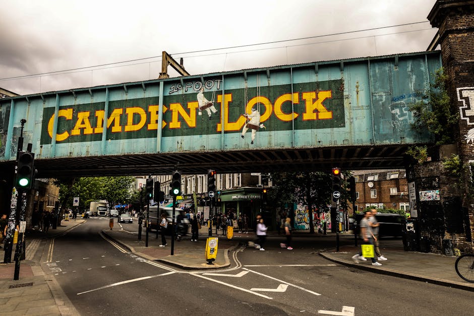 A large, weathered railway bridge with a faded turquoise metal structure spans across a street in Camden Lock, London. The bridge displays a prominent yellow and black 'CAMDEN LOCK' sign painted on its side, with a small illustration of a girl hanging from the letter 'A' in 'CAMDEN'. Below, the street scene features pedestrian crossings with people walking, some carrying bags or backpacks, and others waiting at traffic lights. Vehicles, including cars and a van, are visible on the road, with some waiting at a red light. The sidewalk area shows a mix of brick buildings, street furniture, and graffiti on nearby walls. In the background, there are trees, additional buildings, and street signs. This scene captures a typical busy urban environment where house removals or furniture transport could be coordinated by [COMPANY_NAME] during a home relocation or moving process in the Camden area near Enfield.