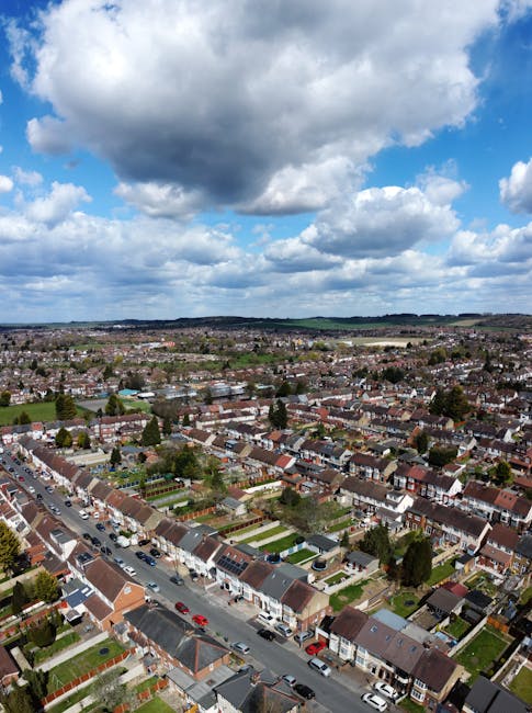 An aerial view of a residential area in Enfield, showing a mix of terraced houses, detached homes, and commercial buildings along a main road. The image includes a courtyard with a few parked cars, a