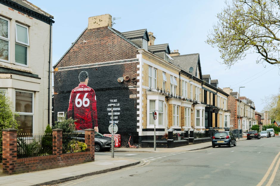 A residential street with a row of terraced houses, including a building with a prominent mural of a person seen from behind wearing a red Liverpool football jersey with the number 66 and the name Alexander-Arnold. The mural is painted on the black brick wall of the house, which has a white cornice and several windows with white frames. The street has parked cars along the curb, and a small tree with fresh green leaves is visible on the right side of the image. A metal pole with a traffic sign stands near the corner, and a pavement runs along the front of the houses. The scene is set in daylight, with an overcast sky. This image captures elements relevant to the home relocation process, such as exterior furniture and street parking, which are considered in planning house removals and furniture transport, as conducted by Removal Companies Enfield.